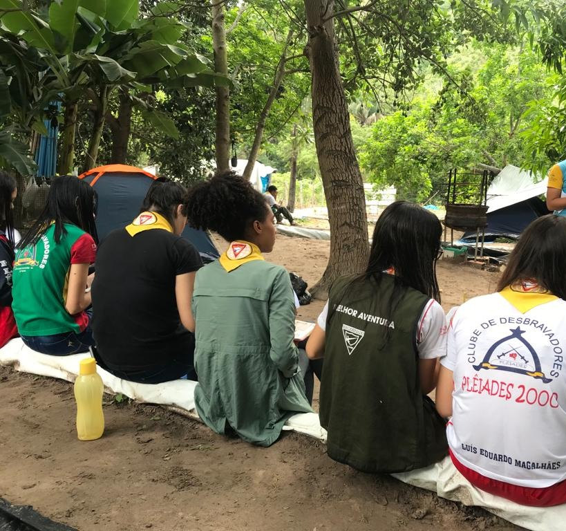 Desbravadoras sentadas de costas sob as árvores, uma delas com a camisa Plêiades 2000
